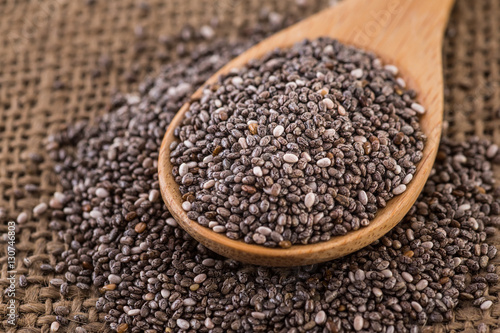 Wooden bowl full with chia seeds and a spoon next to it on sack cloth background.