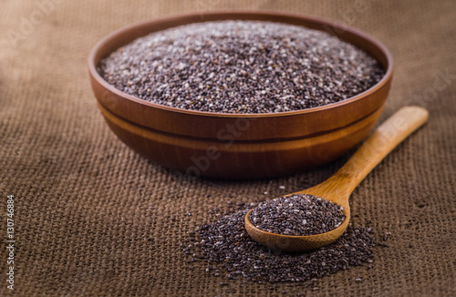 Wooden bowl full with chia seeds and a spoon next to it on sack cloth background.