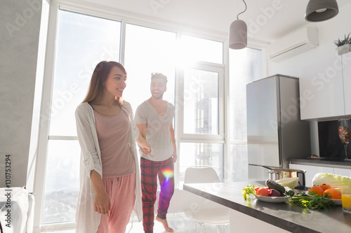 Young Couple Holding Hands In Kitchen, Asian Woman Leading Hispanic Man Modern Apartment With Big Windows Interior © mast3r