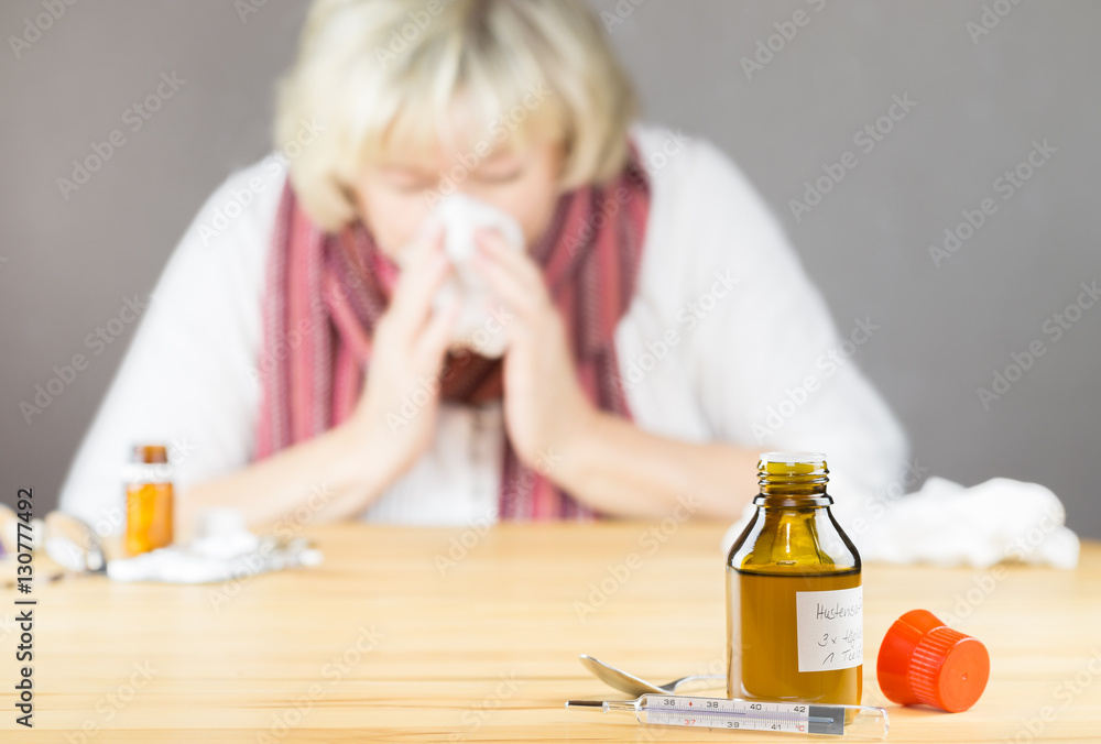 Frau mit Schnupfen und Grippe, im Vordergrund Hustensaft und Fieberthermometer Frau mit Schnupfen und Grippe, im Vordergrund Hustensaft und Fieberthermometer