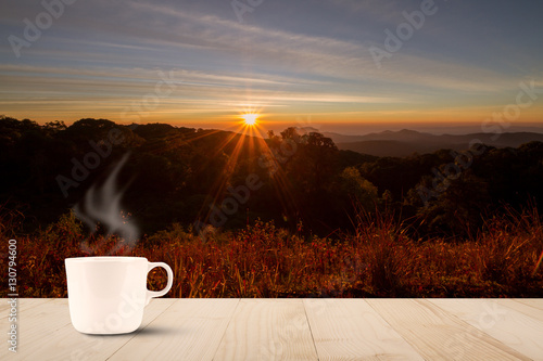 Hot coffee cup with steam on wooden table top on blurred meadow and mountain with sunlight with flare during sunrise