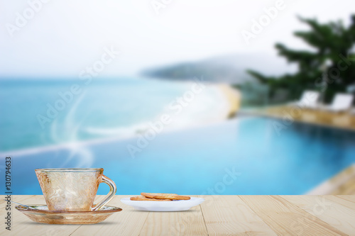 Hot coffee with steam on wooden table top on blurred pool and beach background