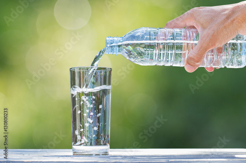 Man's hand holding plastic bottle water and pouring water into glass on wooden table on blurred green bokeh background
