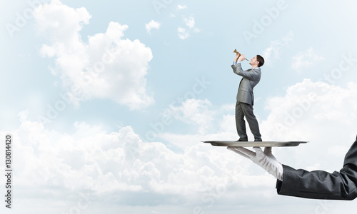 Businessman on metal tray playing fife against blue sky background
