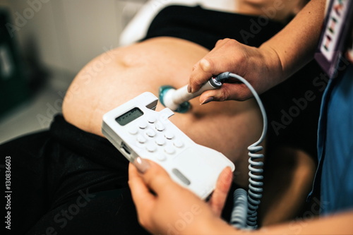 Close up of ultrasound examination of the fetus. Pregnant woman in gynecological clinic. Prenatal testing of young blond woman with hands of doctor. ultrasound scanner in hands of doctor examining her photo