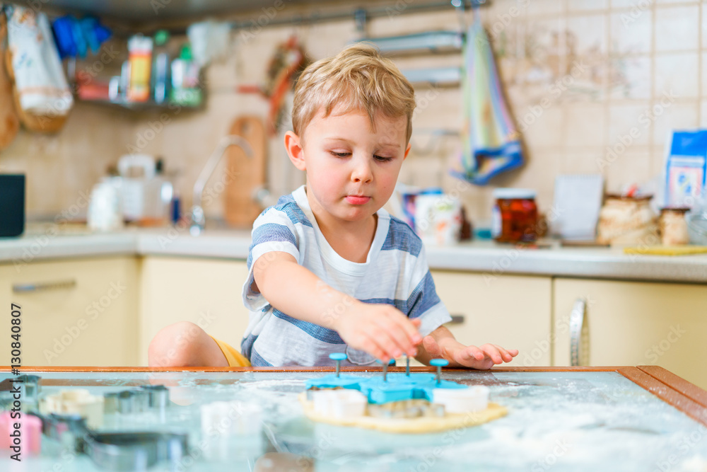 Fototapeta premium Little blond kid plays with molds for making ginger biscuits or cookies, sitting at the kitchen table with raw dough sheet and wheat flour. Protruding lower lip.