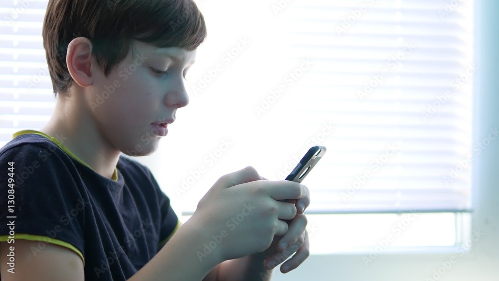 Teenage boy holding a smartphone online games web search social media sitting by the window Teenage boy holding a smartphone online games web search social media sitting by the window