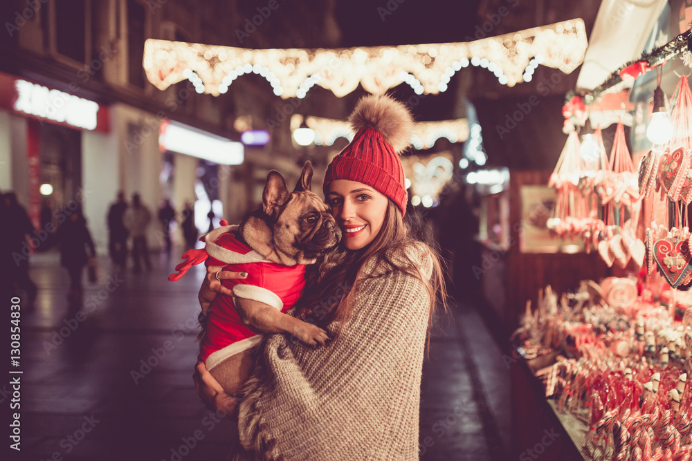 Beautiful young brunette woman with French bulldog enjoying Christmas or New Year night on a city street.