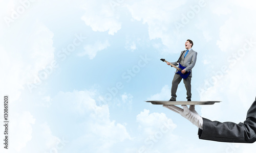 Businessman on metal tray playing electric guitar against blue sky background