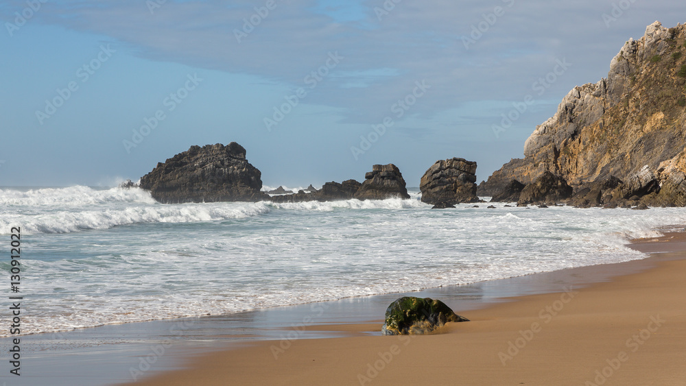 Adraga Beach (Praia da Adraga) in Portugal. Beautiful place, golden sand. Rough sea, rocks, whitewash, blue sky.