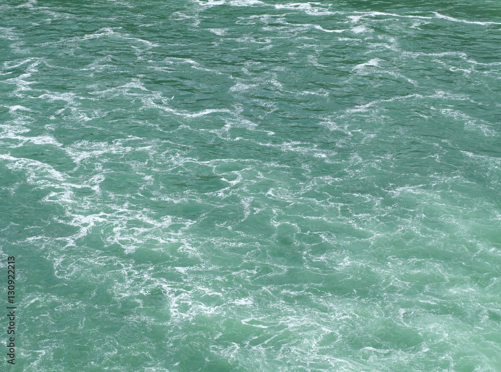 Naturally emerald colored clean and clear water surface with light but very foamy waves flowing down a river after a waterfall. The foam creates a marble looking pattern.