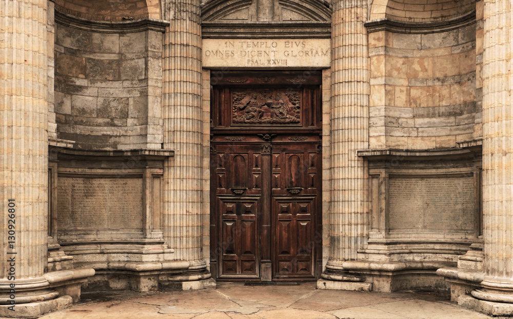 Heavy wooden double door outside of an old church. Circle wall with square stones and pillars. Latin inscriptions on the sides and the arch. Religious symbolic relief on top. Rectangle door frame. Heavy wooden double door outside of an old church. Circle wall with square stones and pillars. Latin inscriptions on the sides and the arch. Religious symbolic relief on top. Rectangle door frame.