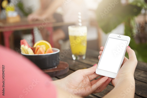 People, technology and leisure concept. Young female making video call to her friend on smart phone, using free wireless internet connection at coffee shop, sitting at table with cocktail and fruits