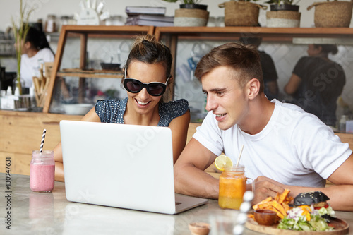 Handsome man and pretty woman in trendy sunglasses smiling while making video call to their friend using free wi-fi on generic laptop pc  having meal  sitting at table in modern cafe interior
