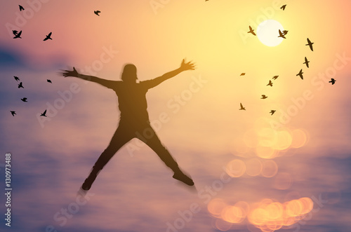 Freedom and feel good concept. Copy space of silhouette happy man jumping on blur tropical sunset beach with birds fly abstract background.
