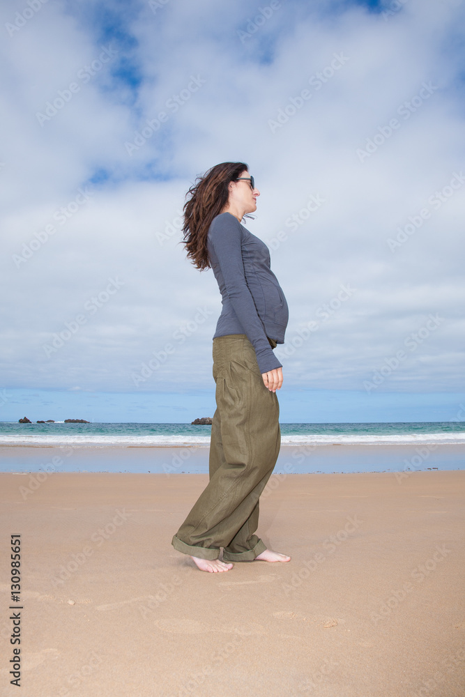 brunette brown hair pregnant woman with grey jersey green trousers and black sunglasses walking in sand beach ocean in Asturias Spain
