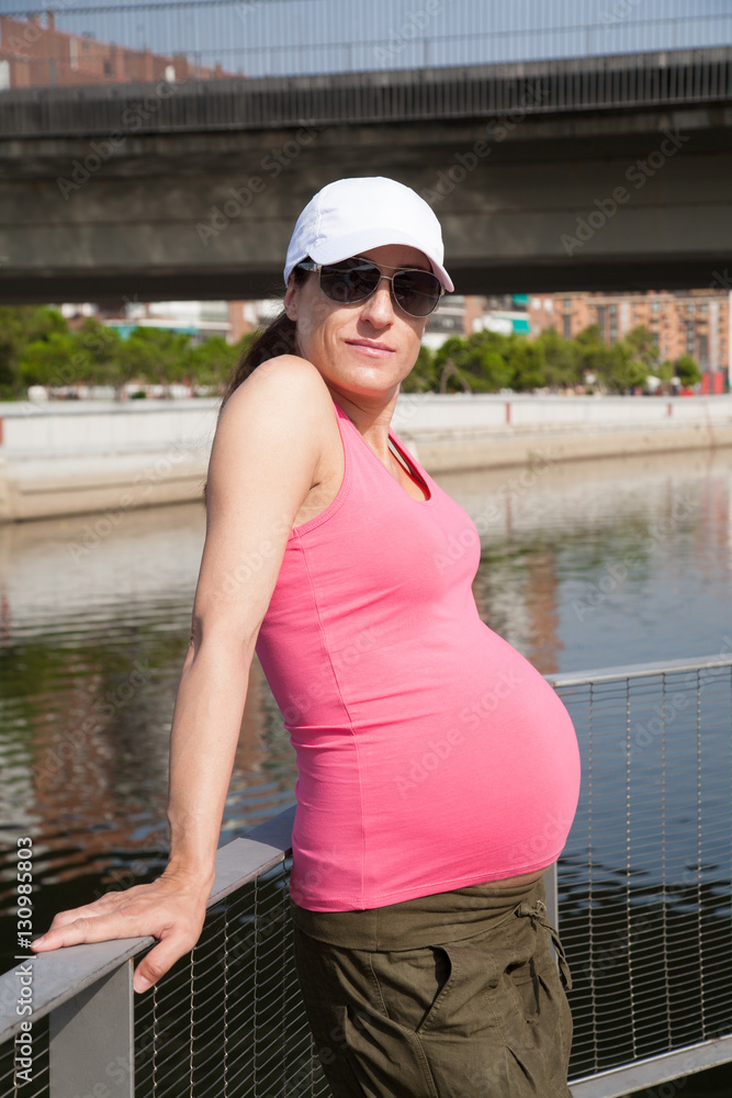 portrait of pregnant woman with white cap pink shirt green trousers and sunglasses in summer next to river in Madrid city
