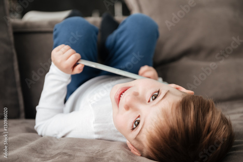 Happy little boy holding tablet computer while lies on sofa