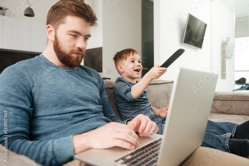 Bearded father using laptop computer while his son watching TV. photo