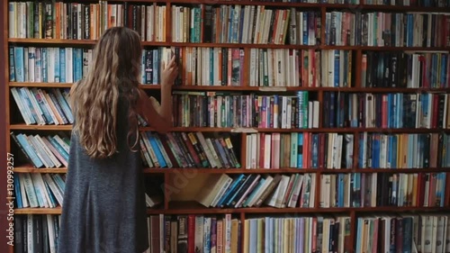 Beautiful young girl with book in hand walks along the bookshelves