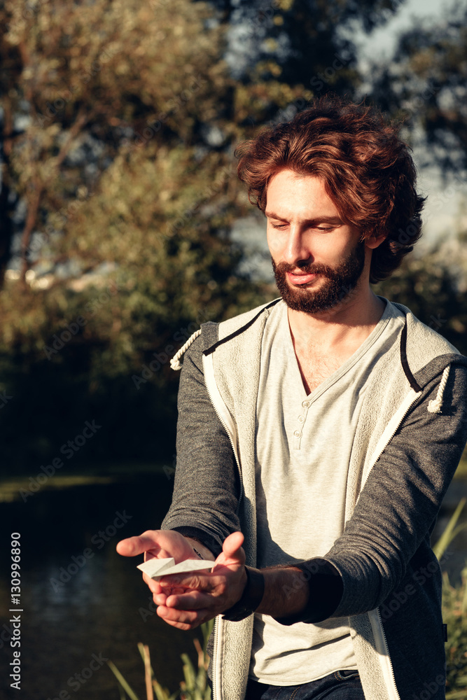 Attractive bearded man holding paper airplane. Stylish guy disporting outdoor with handmade origami plane, nature background. Rest, relax, open-air game, leisure, pastime concept Attractive bearded man holding paper airplane. Stylish guy disporting outdoor with handmade origami plane, nature background. Rest, relax, open-air game, leisure, pastime concept