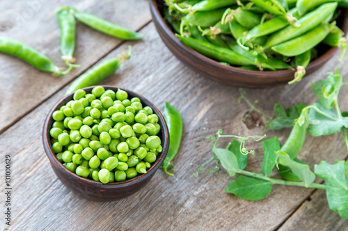 young peas in a clay plate on a wooden table. standing next to a plate of pea pods. next to the plates is the stalk of peas.