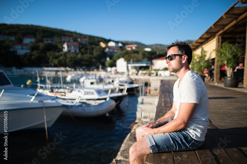 Young man sitting on wooden pier, holding glass of wine