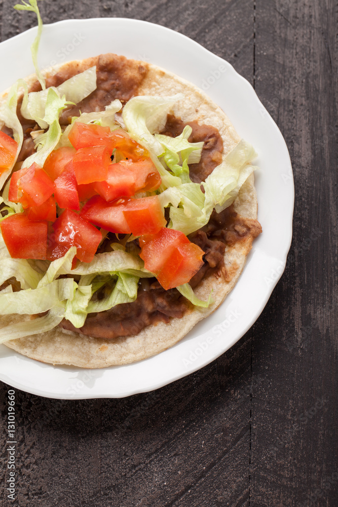 Corn tortilla tostada with refried beans, lettuce, salsa, and tomatoes top view on a dark wooden background Corn tortilla tostada with refried beans, lettuce, salsa, and tomatoes top view on a dark wooden background