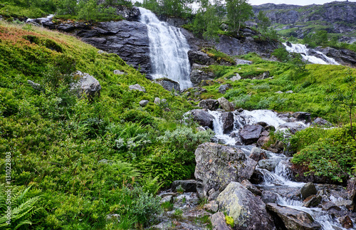 Green and luxuriant nature in Norway, with gushing water in waterfalls and calmly running water on terraced  boulders, beneath the high mountain ridge