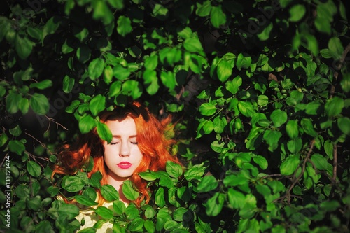 Head of a young attractive girl with closed eyes and flaming red hair peeking through the green leaves of tree