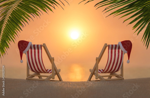 Two deckchairs with red Christmas Santa hats for romantic couple against sunset under the palm