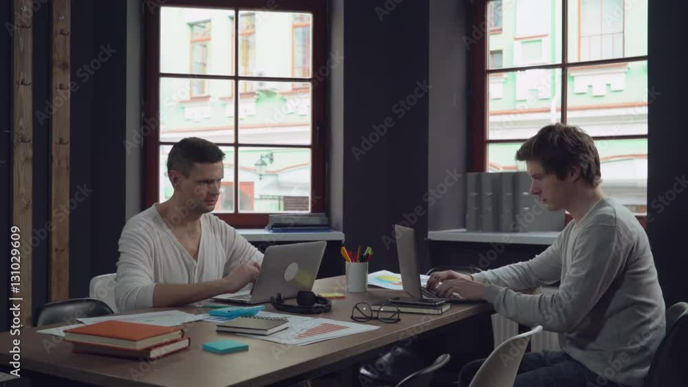 Two young businessmen working in the modern contemporary office with big windows. Group of people works with computer sitting at the wooden desk with paper documents. Happy startup team. Two young businessmen working in the modern contemporary office with big windows. Group of people works with computer sitting at the wooden desk with paper documents. Happy startup team.