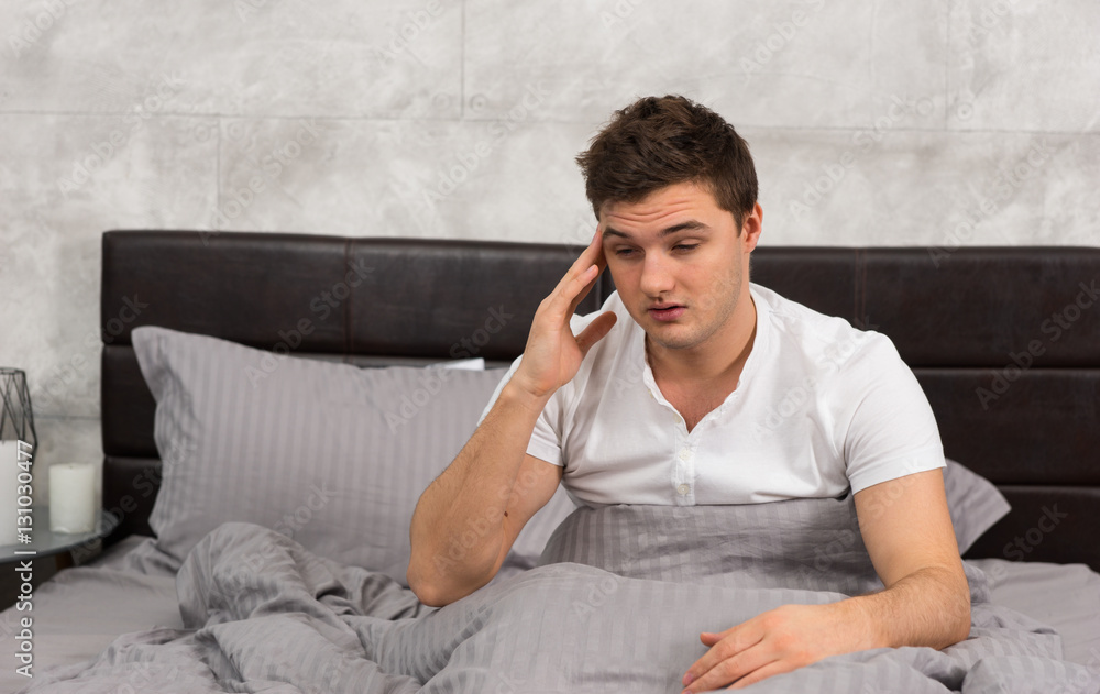 Young man woke up with hangover sitting in stylish bed with grey Young man woke up with hangover sitting in stylish bed with grey