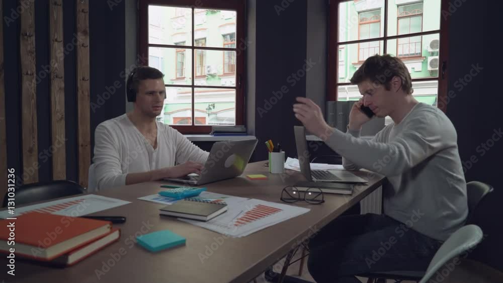 Two young man working in the office. Handsome caucasian man talking on the smartphone. guy with black hair listening music in headphones sitting at the working place using laptop. Startup team use Two young man working in the office. Handsome caucasian man talking on the smartphone. guy with black hair listening music in headphones sitting at the working place using laptop. Startup team use
