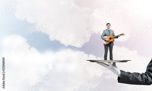 Businessman on metal tray playing acoustic guitar against blue sky background