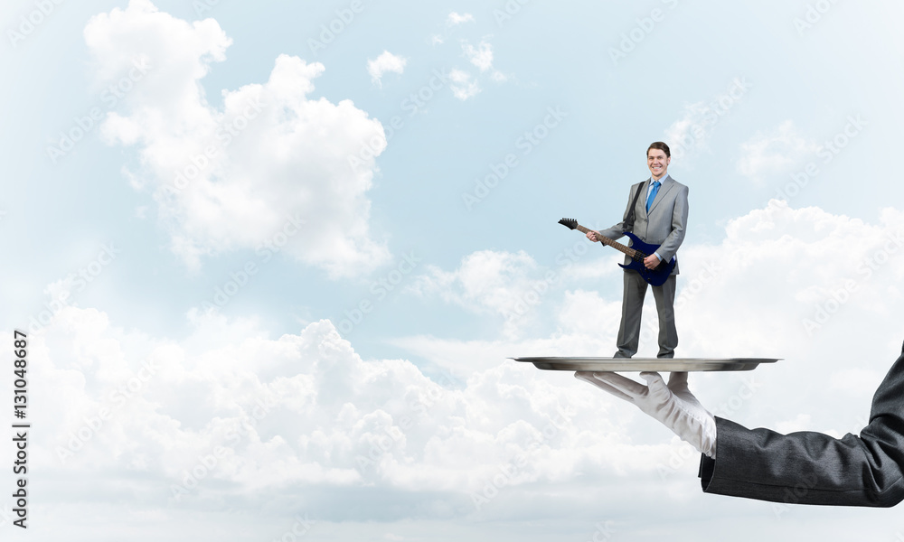 Businessman on metal tray playing electric guitar against blue sky background Businessman on metal tray playing electric guitar against blue sky background