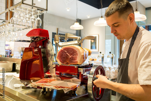 young European Italian chef slicing cutting parma ham cure meat Iberia on red collar slicer commercial kitchen photo