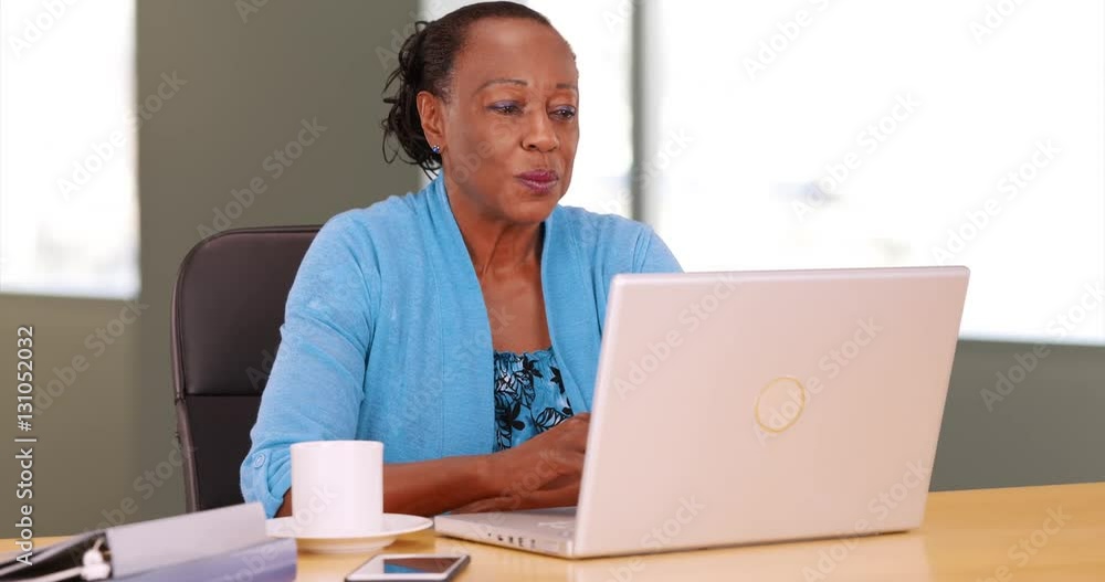 A black businesswoman happily uses her laptop at her desk. An African American woman works on her computer A black businesswoman happily uses her laptop at her desk. An African American woman works on her computer