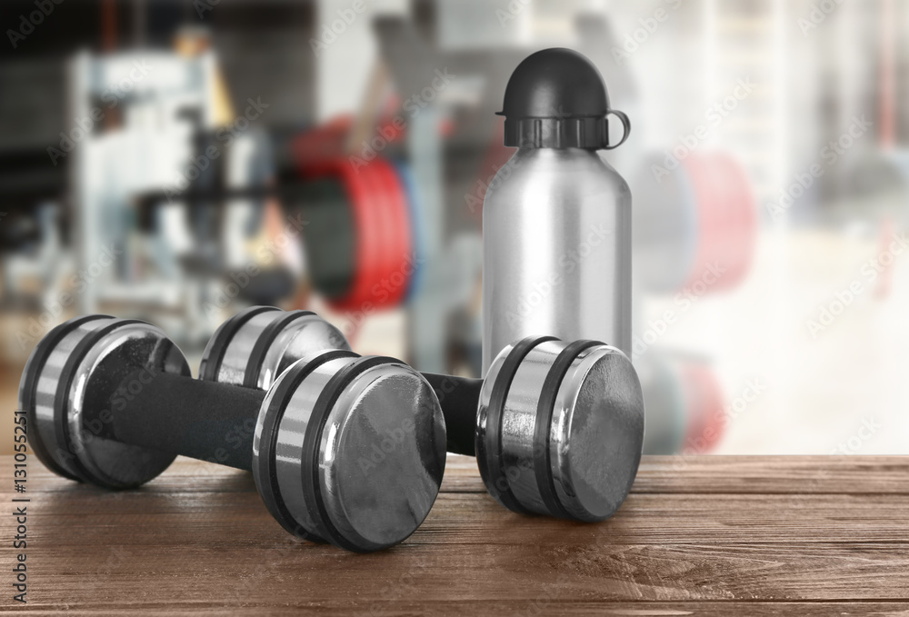 Dumbbells with bottle on table against blurred gym interior background Dumbbells with bottle on table against blurred gym interior background