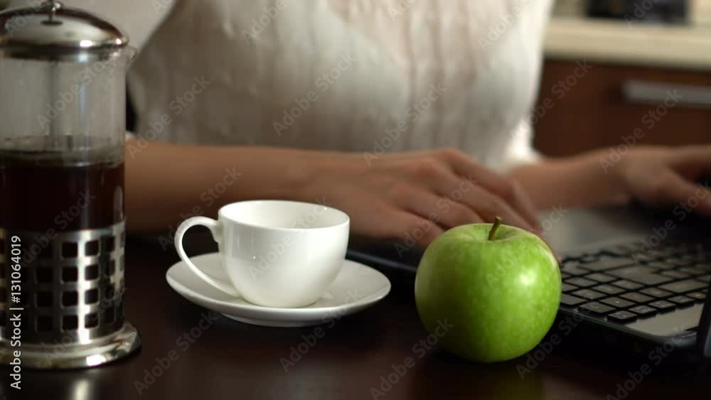 Beautiful young woman using laptop at home in the kitchen. Pouring a cup of coffee. Apple on table. Beautiful young woman using laptop at home in the kitchen. Pouring a cup of coffee. Apple on table.