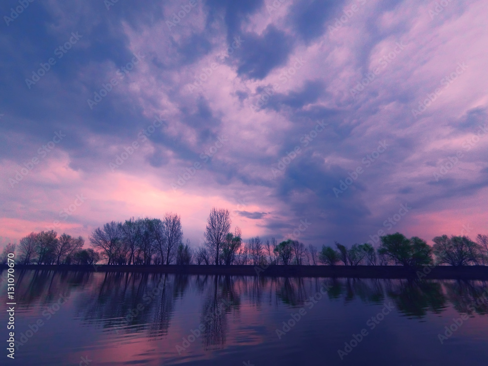 Picturesque view of the river with reflections of the trees of far riverbank. Dramatic evening scene under stormy clouds. Volga-river near Astrakhan, Russia Picturesque view of the river with reflections of the trees of far riverbank. Dramatic evening scene under stormy clouds. Volga-river near Astrakhan, Russia
