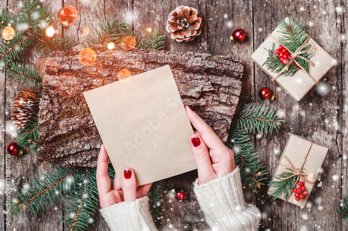 Female hand writing a letter to Santa on wooden background with Christmas gifts, bark texture, Fir branches, pine cones, red decorations. Xmas and Happy New Year composition. Flat lay, top view