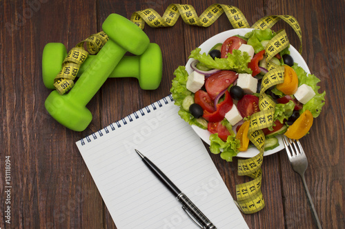 Dumbbell, vegetable salad and measuring tape on rustic wooden table.