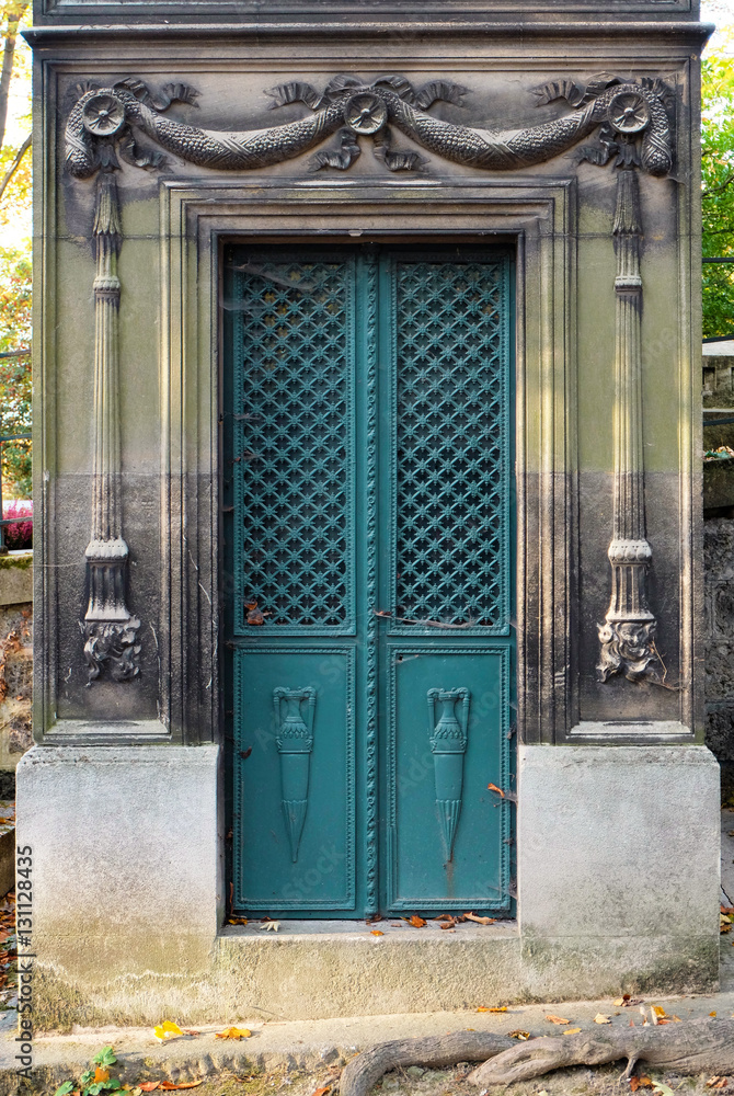 Naklejka premium Rectangular turquise double door made of steel. Ornaments of vases, floral / cross patterns. The ornate chiseled stone door frame contains carved reliefs of torches. An old tomb / crypt at a graveyard
