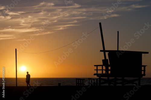 Silhouette of one lone man walking on the beach and one baywatch cabin against the sunset sun and colorful soft gradient in the sky produced by a diffused light, delicate cloud's stripes and the ocean