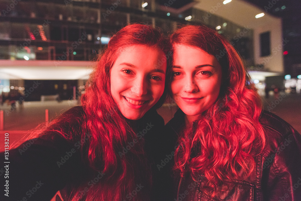 Obraz premium Two young beautiful caucasian women friends outdoor in the city night having fun taking selfie illuminated by red light -social network, sharing, vanity concept