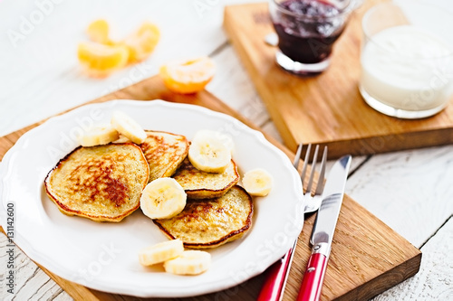 Morning pancakes with banana and coconut on a white plate with red fork and knife. Bright healthy breakfast. White wooden table