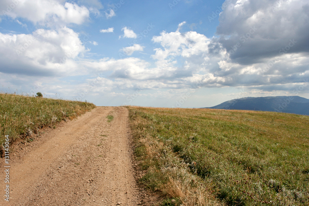 The simple landscape with the earth-road, the distant mountain, the field with green grass and the sky with clouds. This photo was taken in Crimean Mountains, on South Demerdzhi mountain.