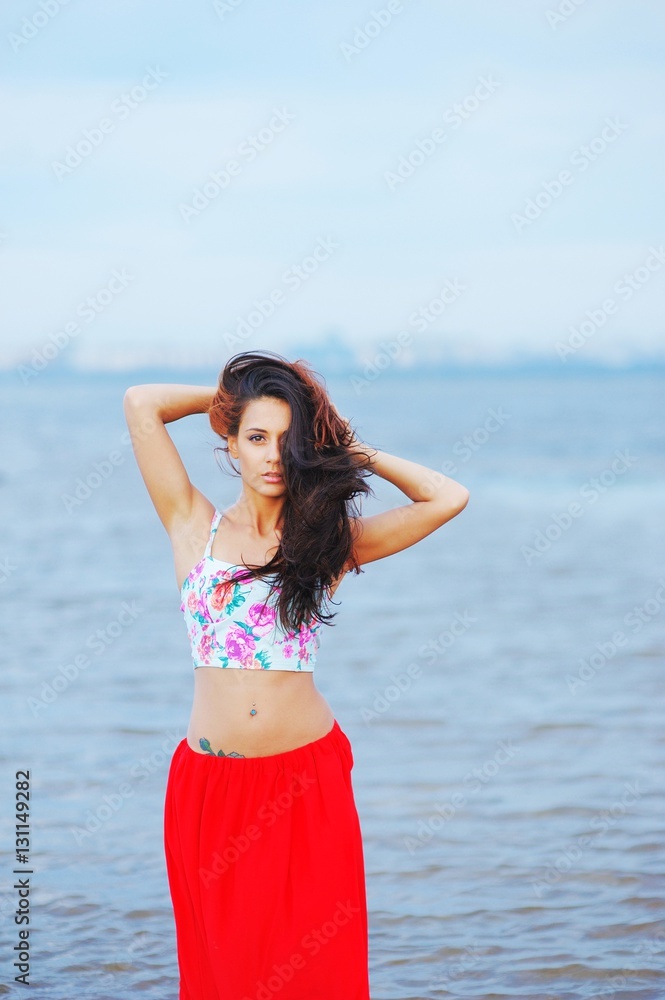 Young pretty long haired brunette girl standing, stretching and relaxing on the shore near the water on the blurred background of the Gulf of Finland, and city's architecture on other side. Young pretty long haired brunette girl standing, stretching and relaxing on the shore near the water on the blurred background of the Gulf of Finland, and city's architecture on other side.