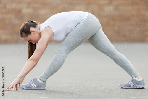 Sporty attractive young woman practicing yoga, standing in Pyramid exercise, Parsvottanasana pose, working out, wearing sportswear, outdoor full length, brick wall background